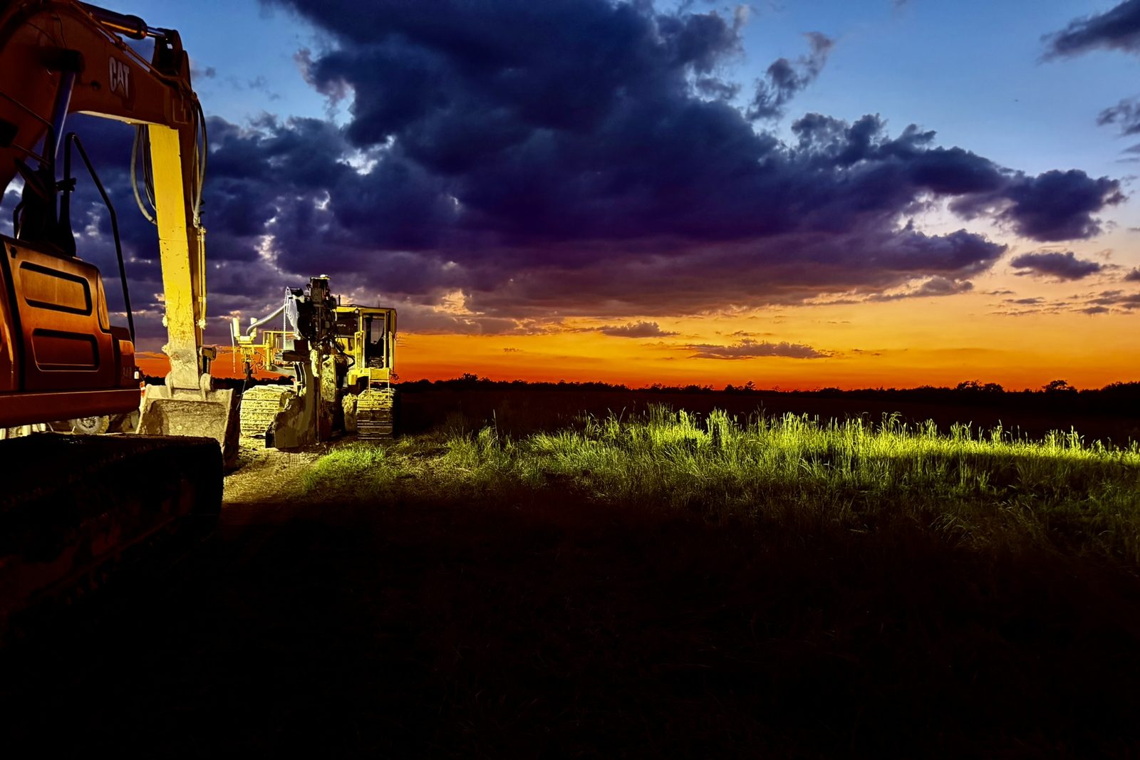 An excavator in the foreground and a tracked trencher beyond it, their work lights on the grass of an open field under a dusk sky.